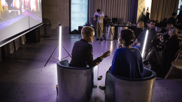 nalevo je plátno, před plátnem stojí neon v prostoru, vepředu  spolu mluví Havránek a  Pachmanová, napravo jsou hosté a poslouchají