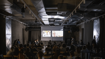 pohled shora čelně na místnost plnou posluchačů, v čele je projekce